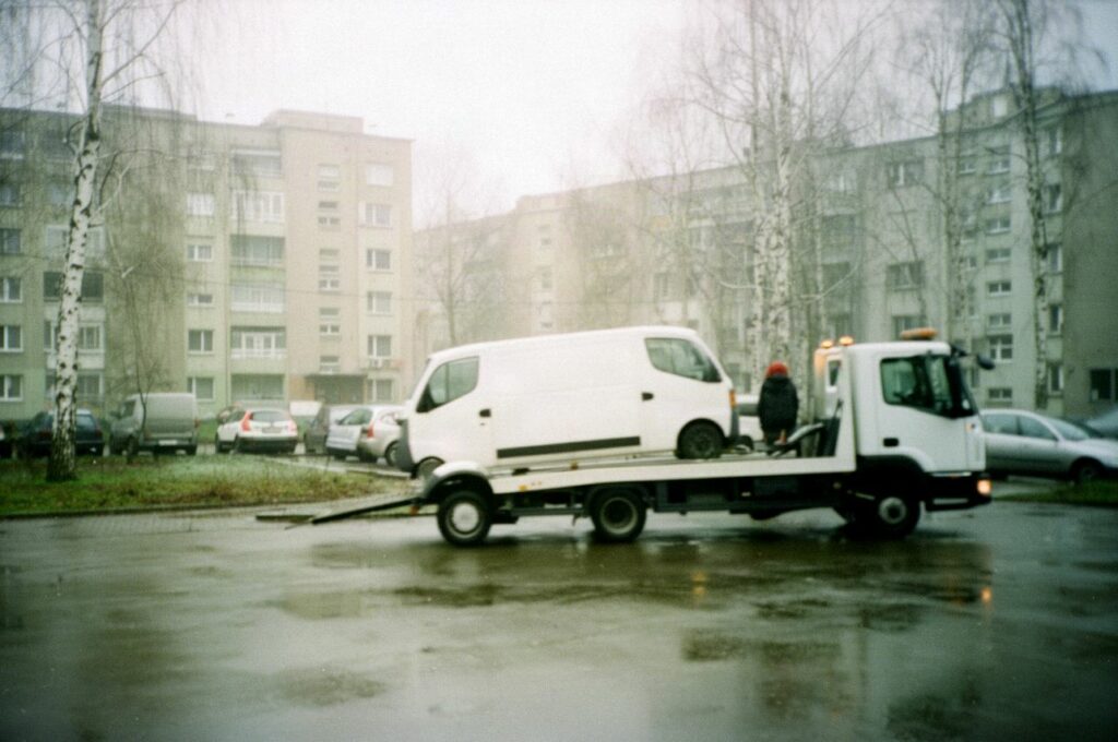 Laweta zabiera biały samochód dostawczy z mokrego parkingu między blokami w pochmurny poranek