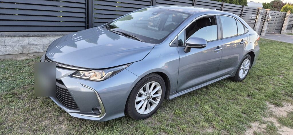 Szara Toyota Corolla sedan na trawie przed ogrodzeniem, widok z przodu i boku, tablica rozmyta