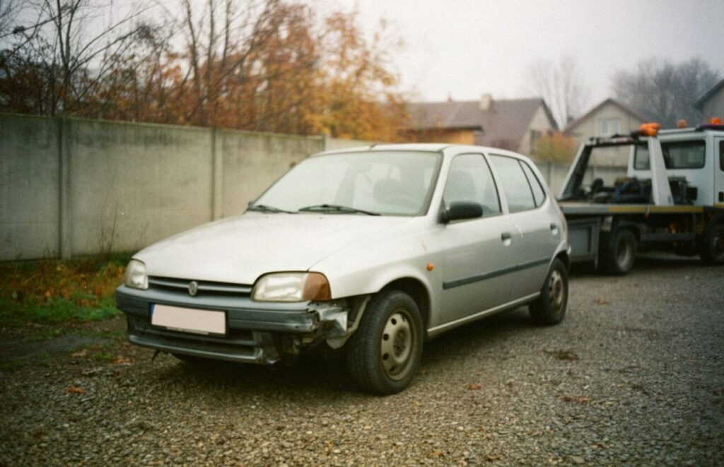 Uszkodzony srebrny samochód osobowy z rozbitym przodem na żwirowym placu, w tle laweta